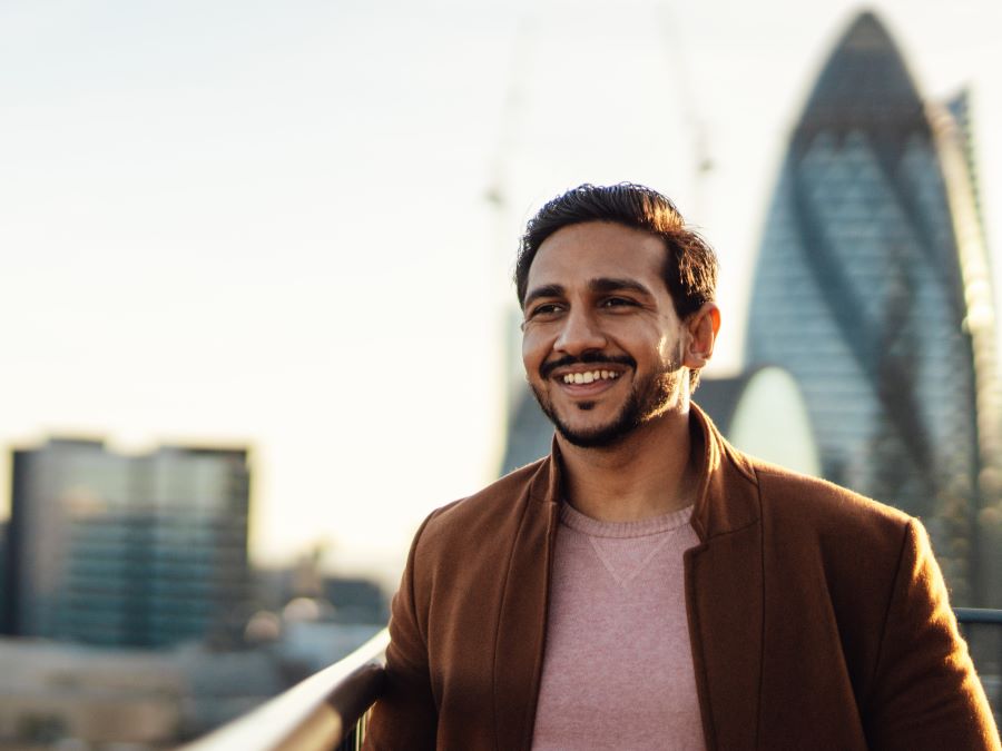A man wearing brown jacket standing on a balcony with London skyline in the background