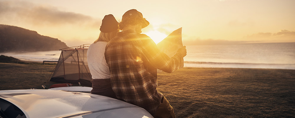 Couple looking at map, sitting on the bonnet of a car, by the ocean