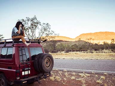 Woman photographing while sitting on car roof at desert by Alice Springs, Australia